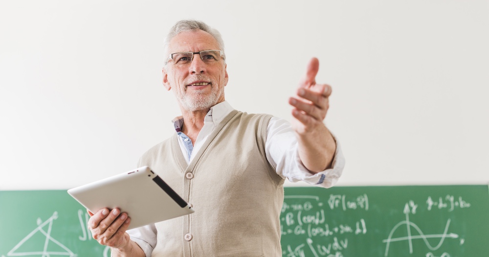 Professor Teaching Class in Front of Blackboard Holding Books and Glasses